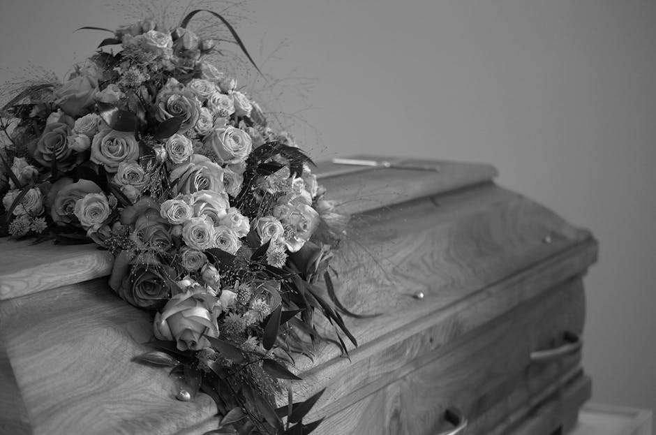 A large, elegant funeral bouquet resting on a polished wooden casket, composed of numerous roses in various shades, predominantly light-colored and dark-colored, with smaller filler flowers and dark g