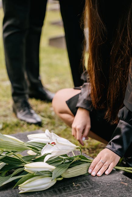A person dressed in black, kneeling on the grass and arranging a bouquet of white lilies with long green leaves, which are laid out on a dark stone surface. The lilies have large, trumpet-shaped petal