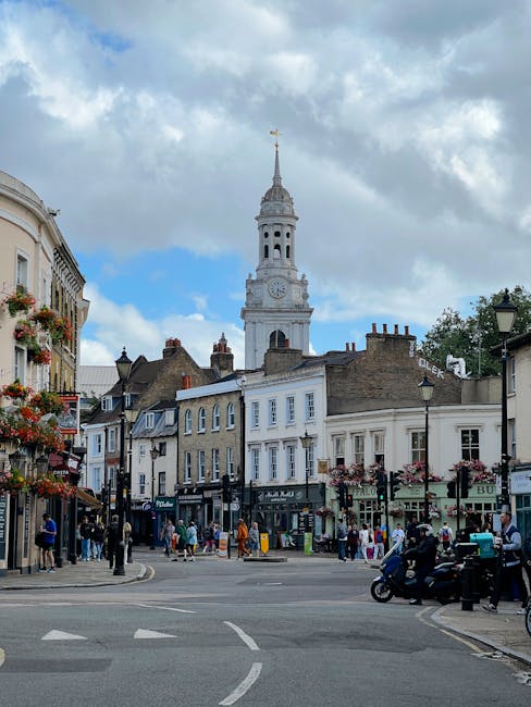 A wide street scene in Tooting with a prominent white clock tower featuring a spire and golden weather vane, set against a partly cloudy sky. Surrounding the square are historic buildings with shopfro