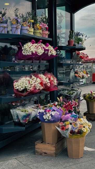 A variety of floral arrangements displayed on blue metal shelves and wooden crates outside a flower shop. The selection includes bouquets of white daisies, pink lilies, purple and yellow mixed flowers