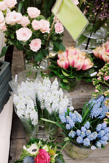 A floral display featuring various fresh flowers arranged in glass vases on a rustic wooden surface. There are light pink roses with lush green leaves in a tall vase, alongside a bouquet of pink and y